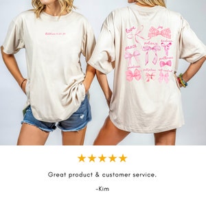 May include: Beige t-shirt with pink text and bow designs. The front of the shirt has the text "Galatians 5:22-23". The back of the shirt features the words "love, peace, joy, kindness, goodness, patience, gentleness, faithfulness, self-control" with bow illustrations.
