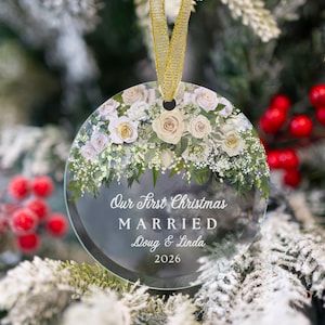 K&ouml;nnte beinhalten: Ein rundes, klares Glasornament mit einem Blumenarrangement aus wei&szlig;en und cremefarbenen Rosen sowie gr&uuml;nem Blattwerk am oberen Rand. Der Text lautet: Our First Christmas MARRIED Doug & Linda 2026. Ein goldenes Band dient zum Aufh&auml;ngen.