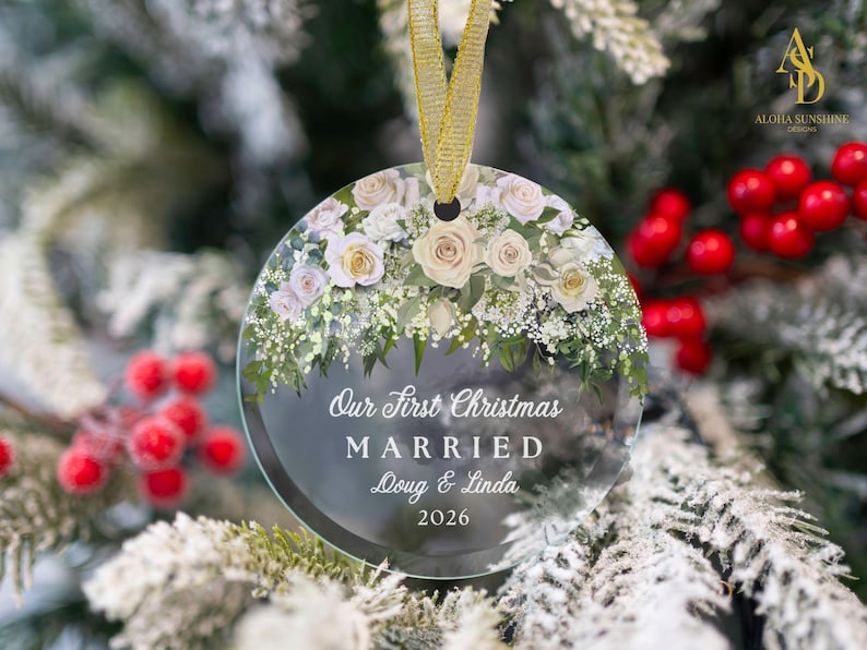 K&ouml;nnte beinhalten: Ein rundes, klares Glasornament mit einem Blumenarrangement aus wei&szlig;en und cremefarbenen Rosen sowie gr&uuml;nem Blattwerk am oberen Rand. Der Text lautet: Our First Christmas MARRIED Doug & Linda 2026. Ein goldenes Band dient zum Aufh&auml;ngen.