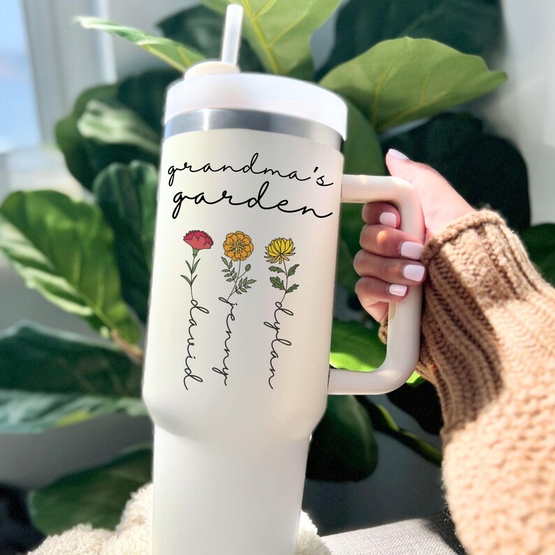 May include: White stainless steel travel mug with a handle and a straw. The mug has a floral design with the text "grandma's garden" and the names "David", "Jenny", and "Dylan" written vertically below the flowers.