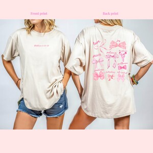 May include: Beige t-shirt with pink text and bow designs. The front of the shirt has the text "Galatians 5:22-23". The back features the words "love, peace, joy, patience, kindness, goodness, gentleness, faithfulness, self control" with bow illustrations.