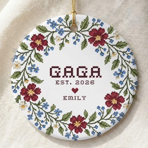 Könnte beinhalten: Ein rundes, weißes Keramikornament mit einem floralen Kranz im Kreuzstich-Stil, bestehend aus roten und blauen Blumen sowie grünen Blättern. Der mittige Text lautet GAGA, EST. 2026, ergänzt durch ein kleines rotes Herz und den Namen EMILY.