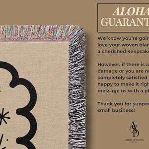 May include: Woven blanket with a beige background and a black floral design. The blanket has a fringed edge with multiple colors. The text "ALOHA GUARANTEE" is in the upper right corner.