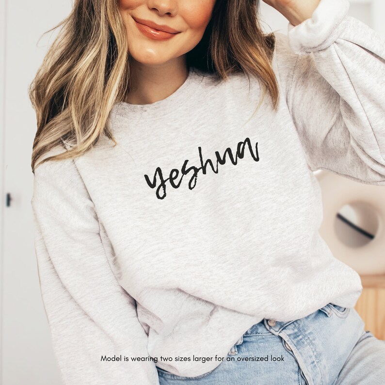 May include: A light gray sweatshirt with the word "yeshua" in black script. The sweatshirt has long sleeves and a crew neck. The model is wearing two sizes larger for an oversized look.