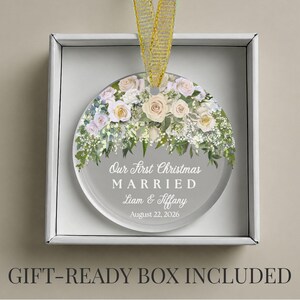 K&ouml;nnte beinhalten: Ein kreisf&ouml;rmiges Glasornament mit einem Blumenarrangement aus wei&szlig;en und blassrosa Rosen sowie gr&uuml;nem Blattwerk. Der Text lautet Our First Christmas MARRIED Liam & Tiffany August 22, 2026. Ein goldenes Band ist befestigt, in einer wei&szlig;en Schachtel.
