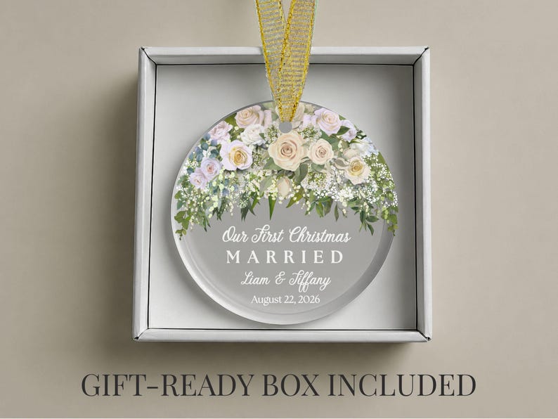 K&ouml;nnte beinhalten: Ein kreisf&ouml;rmiges Glasornament mit einem Blumenarrangement aus wei&szlig;en und blassrosa Rosen sowie gr&uuml;nem Blattwerk. Der Text lautet Our First Christmas MARRIED Liam & Tiffany August 22, 2026. Ein goldenes Band ist befestigt, in einer wei&szlig;en Schachtel.