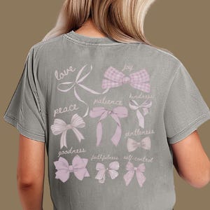 May include: Grey t-shirt with a design of pink bows and words such as "love", "peace", and "joy". The shirt has a vintage, faded appearance.