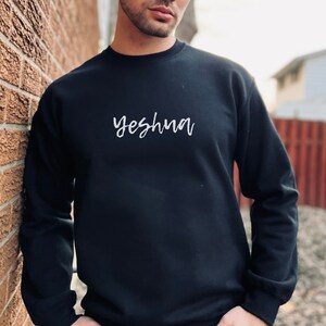 May include: Black crew neck sweatshirt with the word "Yeshua" in white script across the chest. The sweatshirt is a solid color and appears to be made of a soft material. The person wearing the sweatshirt is leaning against a brick wall.
