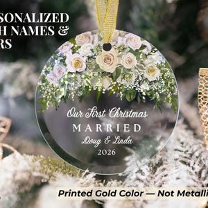 K&ouml;nnte beinhalten: Ein runder, durchsichtiger Glasanh&auml;nger mit einem Blumenarrangement aus wei&szlig;en und blassrosa Rosen sowie gr&uuml;nem Blattwerk am oberen Rand. Der Text lautet Our First Christmas MARRIED Doug & Linda 2026. Er h&auml;ngt an einem goldenen Band.