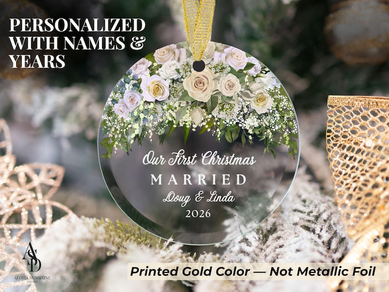 K&ouml;nnte beinhalten: Ein runder, durchsichtiger Glasanh&auml;nger mit einem Blumenarrangement aus wei&szlig;en und blassrosa Rosen sowie gr&uuml;nem Blattwerk am oberen Rand. Der Text lautet Our First Christmas MARRIED Doug & Linda 2026. Er h&auml;ngt an einem goldenen Band.