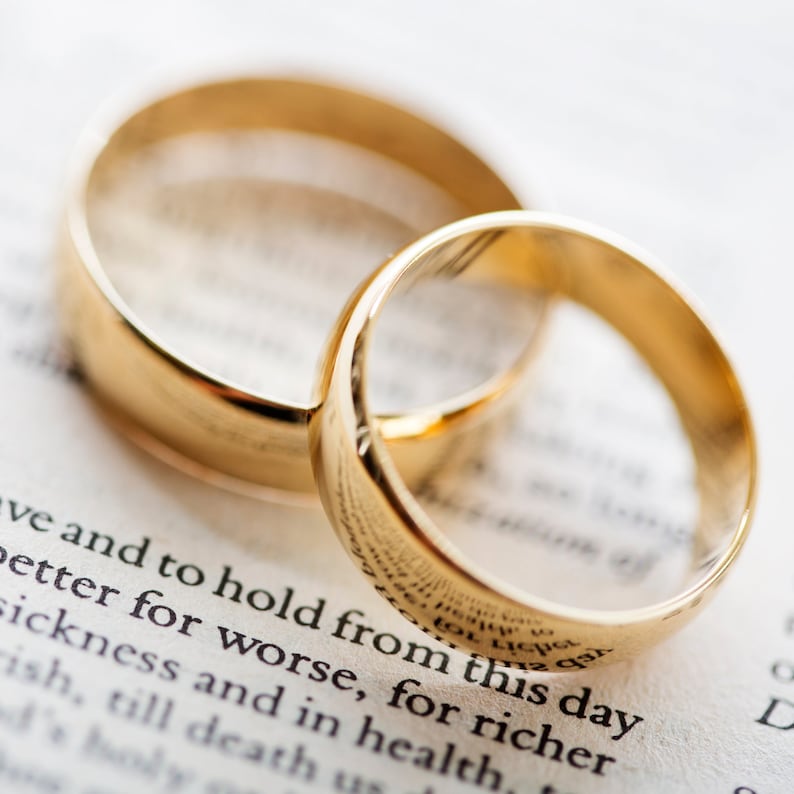 May include: Two gold wedding bands, one slightly larger than the other, are lying on a white page with text. The text reads "Love and to hold from this day better for worse, for richer sickness and in health, till death us"