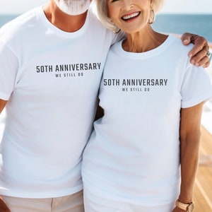 May include: A couple wearing white t-shirts with the text "50th Anniversary We Still Do" printed on them. They are standing close together and smiling.
