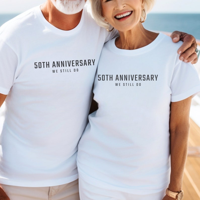 May include: A couple wearing white t-shirts with the text "50th Anniversary We Still Do" printed on them. They are standing close together and smiling.