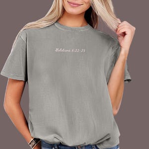 May include: A grey short-sleeved t-shirt with the text "Galatians 5:22-23" in a light pink script. The shirt is a solid colour and has a crew neck.