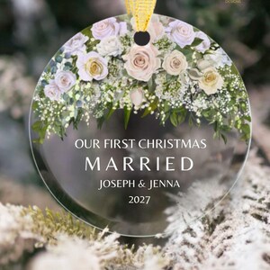K&ouml;nnte beinhalten: Ein rundes, transparentes Acryl-Ornament mit einem Blumenarrangement aus blassrosa und wei&szlig;en Rosen sowie gr&uuml;nem Blattwerk am oberen Rand. Der Text lautet OUR FIRST CHRISTMAS MARRIED JOSEPH & JENNA 2027. Ein goldenes Band ist oben befestigt.