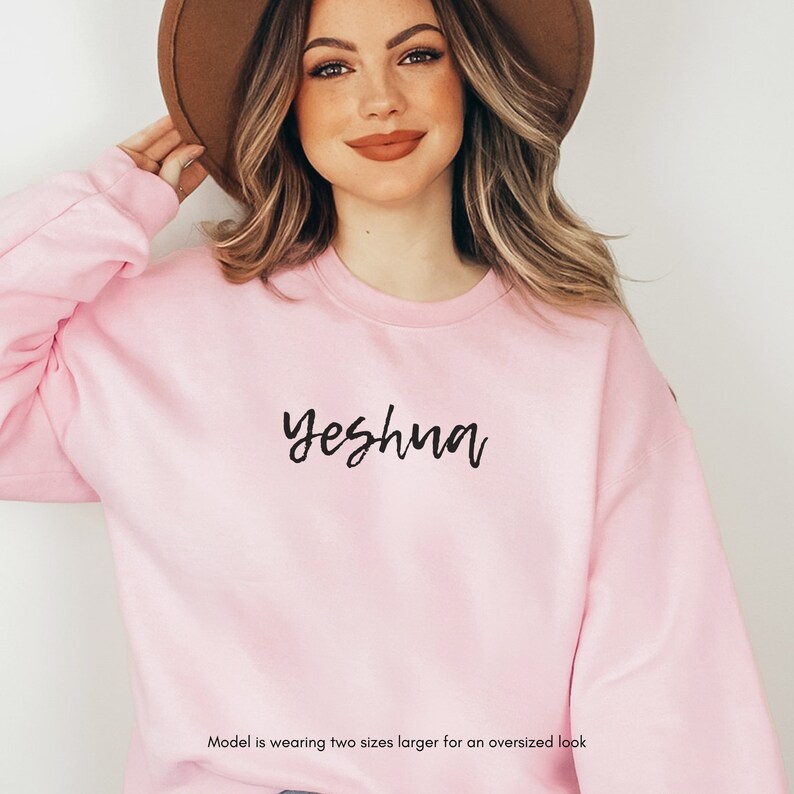 May include: A light pink sweatshirt with the word "Yeshua" in black script. The sweatshirt has long sleeves and a crew neck. The model is wearing two sizes larger for an oversized look.