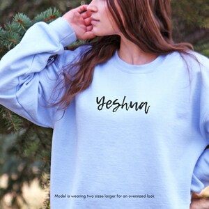 May include: A light blue sweatshirt with the word "Yeshua" in black script. The sweatshirt is a solid color and has a crew neck. The model is wearing two sizes larger for an oversized look.
