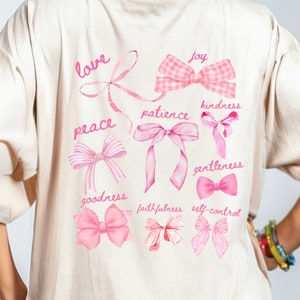 May include: A cream-coloured t-shirt featuring a pink bow design and inspirational words such as "love", "peace", and "joy". The shirt has a watercolour style with the text "Aloha Sunshine Designs" at the top.