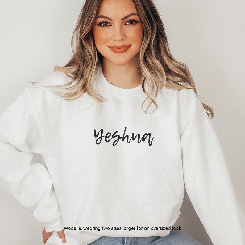 May include: White sweatshirt with the word "Yeshua" in black script across the chest. The sweatshirt is a crew neck style and appears to be made of a soft material. The model is wearing two sizes larger for an oversized look.