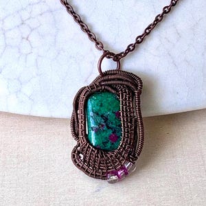 May include: A handcrafted pendant necklace featuring a rectangular green stone with red inclusions, encased in intricately woven copper wire. The pendant hangs from a delicate copper chain. The necklace is displayed on a white surface.