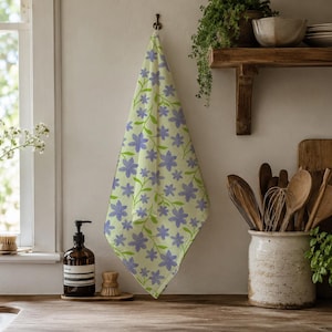 May include: A rectangular kitchen towel featuring a light yellow background with a repeating pattern of purple five-petalled flowers and green stems. The towel is hung vertically, displaying a soft fabric texture and a consistent floral print design.