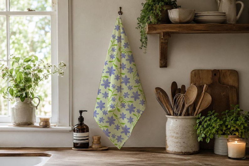 May include: A rectangular kitchen towel featuring a light yellow background with a repeating pattern of purple five-petalled flowers and green stems. The towel is hung vertically, displaying a soft fabric texture and a consistent floral print design.