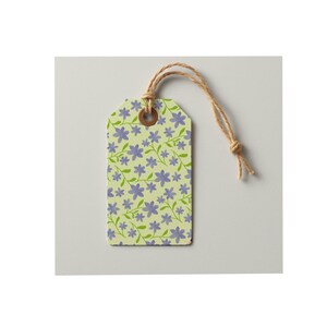 May include: A rectangular paper gift tag featuring a light yellow background with a repeating pattern of small purple flowers and green stems. The tag has a brown reinforced eyelet at the top with a piece of natural tan twine looped through it.
