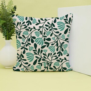Pode incluir: Uma almofada quadrada com fundo creme e um padr&atilde;o repetitivo de folhas verde-escuras e flores estilizadas em tom azul-petr&oacute;leo claro. O design consiste em caules e folhagens interligados que cobrem toda a superf&iacute;cie frontal da almofada.