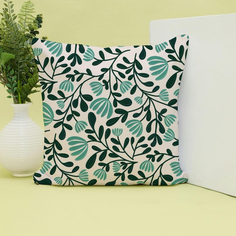 Pode incluir: Uma almofada quadrada com fundo creme e um padr&atilde;o repetitivo de folhas verde-escuras e flores estilizadas em tom azul-petr&oacute;leo claro. O design consiste em caules e folhagens interligados que cobrem toda a superf&iacute;cie frontal da almofada.