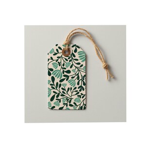 Pode incluir: Uma etiqueta de presente retangular de papel com fundo creme e um padr&atilde;o repetitivo de folhas verde-escuras e formas florais em tom azul-petr&oacute;leo claro. A etiqueta possui um ilh&oacute; refor&ccedil;ado castanho e um cordel bege no orif&iacute;cio superior.