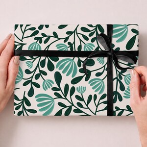 Pode incluir: Uma caixa de presente retangular embrulhada em papel branco com um padr&atilde;o repetitivo de folhas verde-escuras e flores estilizadas em tom azul-petr&oacute;leo claro. O pacote &eacute; amarrado com uma fita de cetim preta fina com um la&ccedil;o &agrave; direita.