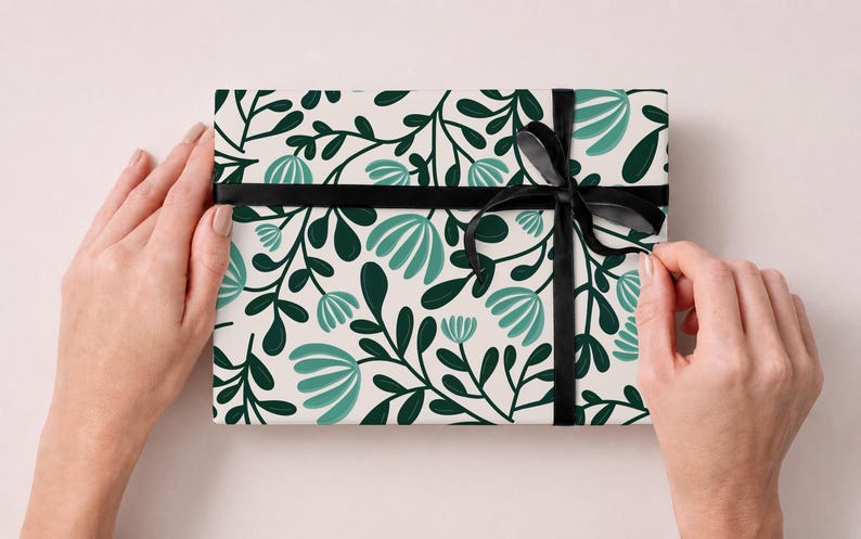 Pode incluir: Uma caixa de presente retangular embrulhada em papel branco com um padr&atilde;o repetitivo de folhas verde-escuras e flores estilizadas em tom azul-petr&oacute;leo claro. O pacote &eacute; amarrado com uma fita de cetim preta fina com um la&ccedil;o &agrave; direita.