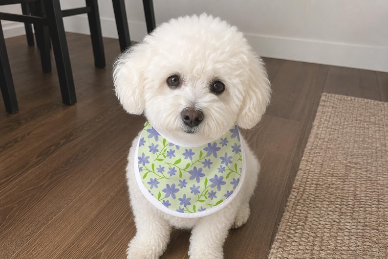 May include: A small white dog with fluffy fur wearing a pet bandana. The bandana features a light green background with a repeating pattern of small purple flowers and thin green stems, finished with a solid white fabric border around the edge.