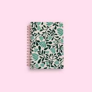 Pode incluir: Um caderno de espiral com uma capa de cor creme apresentando um padr&atilde;o repetitivo de folhas verde-escuras e flores em tom azul-petr&oacute;leo claro. A encaderna&ccedil;&atilde;o consiste numa espiral de arame met&aacute;lico em ouro rosa no lado esquerdo do caderno.
