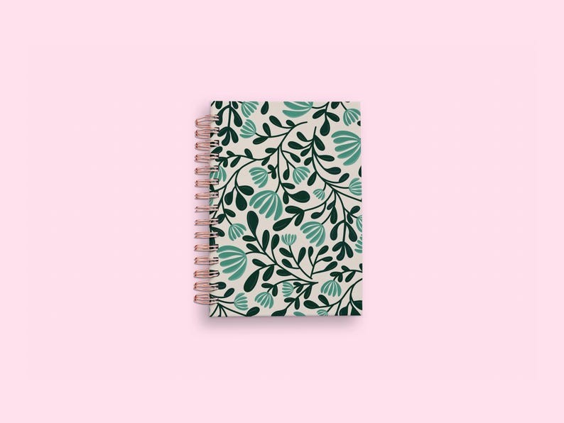 Pode incluir: Um caderno de espiral com uma capa de cor creme apresentando um padr&atilde;o repetitivo de folhas verde-escuras e flores em tom azul-petr&oacute;leo claro. A encaderna&ccedil;&atilde;o consiste numa espiral de arame met&aacute;lico em ouro rosa no lado esquerdo do caderno.