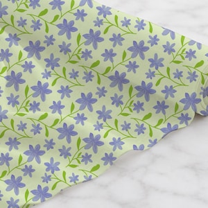 May include: A roll of fabric featuring a repeating pattern of light purple five-petalled flowers with green stems and leaves on a pale lime green background. The fabric is wound around a central cardboard tube, displaying a continuous floral print.