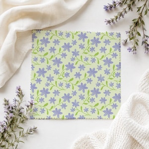 May include: A square fabric swatch featuring a repeating pattern of light purple five-petaled flowers with green stems and leaves on a pale lime green background. The edges of the fabric are finished with a decorative pinked zigzag cut.