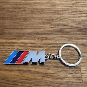 Könnte beinhalten: Ein silberfarbener Metall-Schlüsselanhänger mit dem BMW M-Logo. Das Logo hat ein Design mit blauen, dunkelblauen und roten Streifen. Der Schlüsselanhänger enthält einen Schlüsselring zum Anbringen von Schlüsseln.