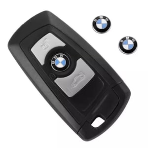 Könnte beinhalten: Schwarzer BMW Autoschlüssel mit silbernen Knöpfen und dem BMW-Logo. Zwei zusätzliche BMW-Logo-Aufkleber sind ebenfalls sichtbar. Der Schlüssel hat Tasten zum Ver- und Entriegeln sowie zum Öffnen des Kofferraums. Der Schlüssel und die Aufkleber befinden sich auf weißem Hintergrund.