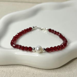 May include: A delicate bracelet featuring faceted red beads and a central white pearl. The bracelet is strung on a silver chain and rests on a white, scalloped dish. A simple and elegant piece of jewelry.