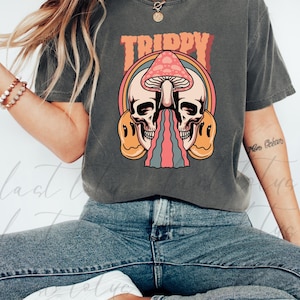 Puede incluir: Camiseta gris oscuro con la palabra "TRIPPY" en naranja sobre un gráfico de calaveras, una seta y un arcoíris. El diseño incluye caras sonrientes y líneas coloridas.