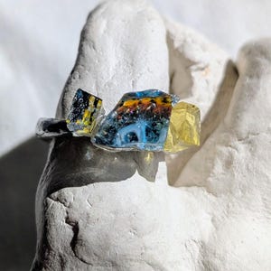 Peut inclure: Une bague unique ornée de fragments de verre colorés dans des tons de bleu, jaune et noir. La monture de la bague est argentée et les morceaux de verre sont de forme irrégulière, créant un design artistique saisissant. La bague est présentée sur une surface blanche texturée.