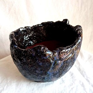 May include: A handmade ceramic bowl with a dark, textured exterior and a deep red interior. The bowl's rim has an irregular, organic shape, and the exterior displays hints of blue and gold.