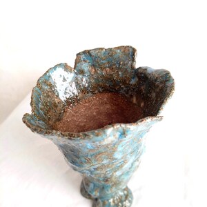 Op de afbeelding: Een handgemaakte keramische vaas met een uniek, getextureerd oppervlak. De vaas heeft een uitlopende, bloembladachtige rand en een voetstuk. Het kleurenpalet omvat tinten blauw, bruin en goud, wat een rustieke, artistieke esthetiek creëert.