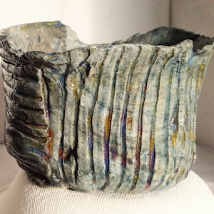 Peut inclure: Pot en céramique fait à la main, de forme carrée et à la surface texturée. Le pot est principalement gris avec des rayures verticales et des touches de bleu, jaune et rouge. Le bord est irrégulier. Posé sur une surface blanche.