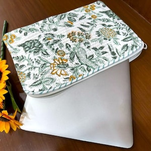 May include: A white laptop sleeve with a floral pattern in green and yellow, with a zipper closure. The sleeve is open, revealing a silver laptop. Two sunflowers are on the left side of the image.