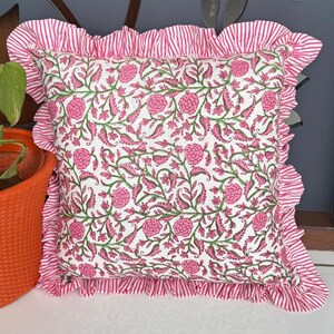 May include: Square decorative pillow with a floral pattern in pink and green on a white background. The pillow has a ruffled edge with pink and white stripes. The pillow is a home decor item.