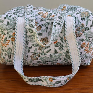 May include: A white cotton tote bag with a floral print in shades of green and orange. The bag has a long handle and a zipper closure.