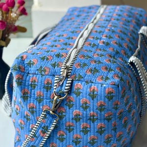 May include: Blue quilted duffel bag with a floral pattern in pink and green. The bag features a white zip and black and white striped straps. It has a brass-coloured metal clasp.