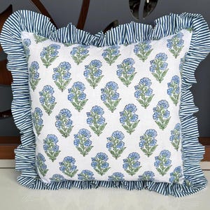 May include: Square throw pillow with a white background and a repeating pattern of blue floral bouquets. The pillow has a ruffled border with blue and white stripes. The pillow is a decorative item for a bed or sofa.